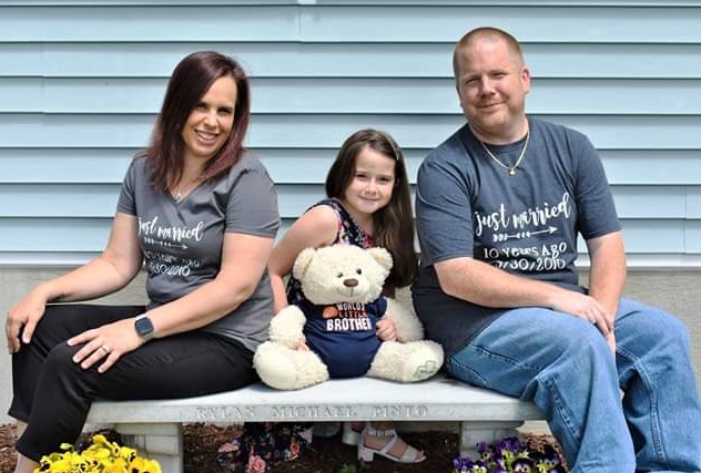 The Pinto Family sitting together. A woman A man and a young girl with a bear siggitng on a bench